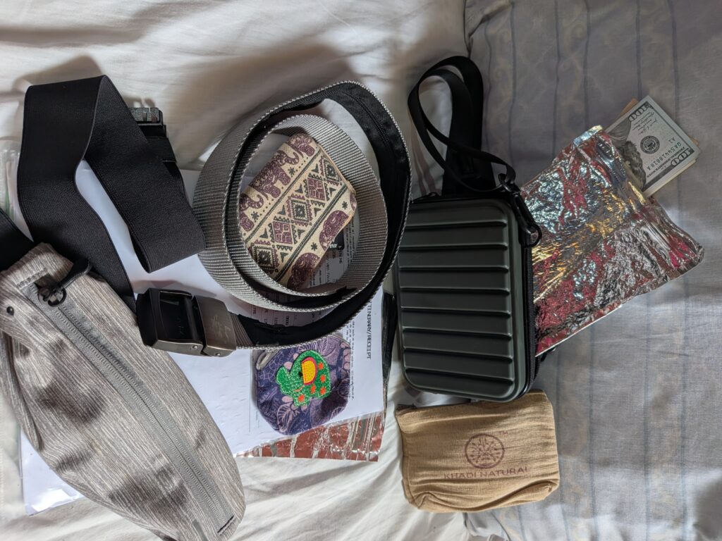 自助旅行的防盜與日常錢包裝備，包括：一個綠色 Travel Blue 親膚烤漆斜背包、一個卡其色布質日用小包、一條灰色的防盜皮帶、一個紫色的零錢包、一個疊起來的格紋鈔票夾、一個藏錢腰包，以及在斜背包上方露出的百元美金紙鈔與銀色夾鏈袋（垃圾袋錢包）。